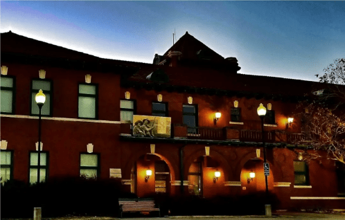 Historic red brick building with arched windows and a decorative roof, illuminated by street lamps at dusk.