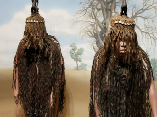 Two figures with long, textured hair and traditional attire stand against a backdrop of a barren landscape.