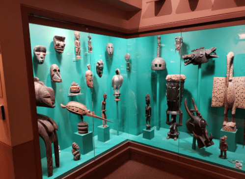 Display of various African sculptures and masks in a glass case, featuring intricate designs and diverse cultural artifacts.