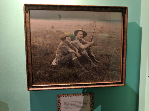 A vintage photo of two people sitting in a grassy field, both wearing hats and holding a fishing rod.