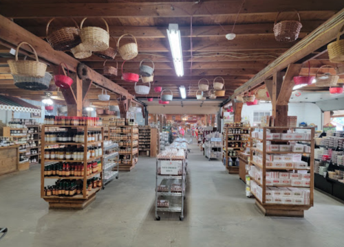 A spacious market interior with wooden beams, shelves of jars and bottles, and hanging baskets overhead.