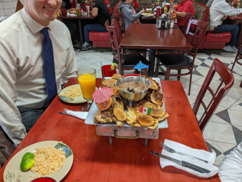 A table at a restaurant with a man smiling, a colorful dish of food with umbrellas, and drinks in a lively setting.