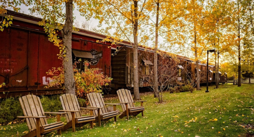 A row of wooden chairs in front of colorful autumn trees and vintage train cars.
