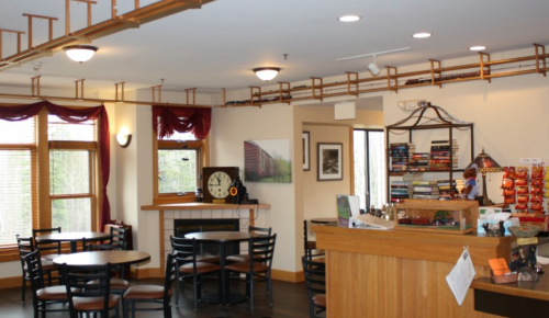 Cozy café interior with wooden furniture, a counter, bookshelves, and large windows letting in natural light.