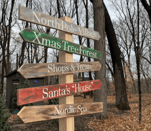 Wooden signpost with directional arrows for North Pole Village, Xmas Tree Forest, Shops & Treats, Santa's "House," and Nordic Spa.