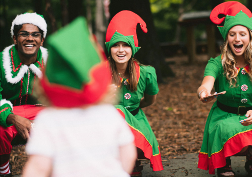 Three cheerful elves in festive outfits smile and interact with a child in a playful outdoor setting.