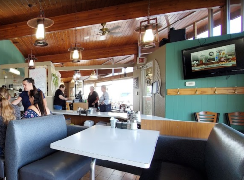 A cozy diner interior with wooden beams, tables, and a TV, featuring staff and customers enjoying their meals.