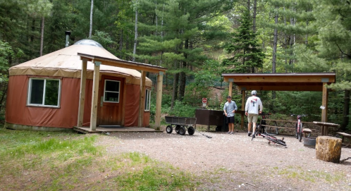 A yurt in a forested area with two people near a picnic table and a bike, surrounded by trees.