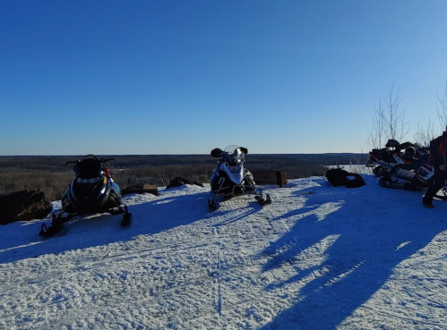 Two snowmobiles parked on a snowy hilltop with a clear blue sky and distant landscape in the background.