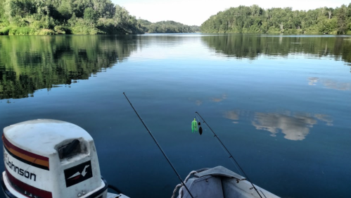 A calm lake scene from a boat, featuring fishing rods and a serene reflection of trees and sky on the water.