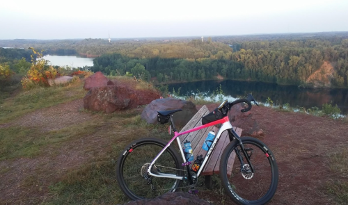A bike rests on a rocky overlook with a scenic view of a river and forested hills in the background during sunset.