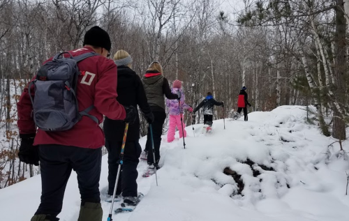 A group of people snowshoeing through a snowy forest trail, surrounded by trees.