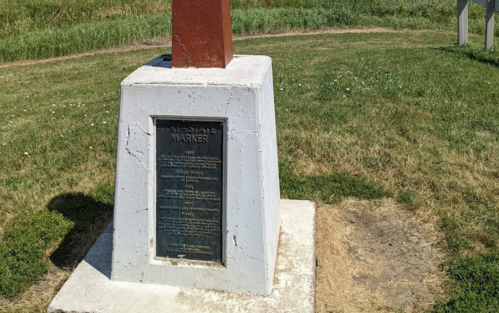 A historical marker on a grassy area, featuring a plaque mounted on a white pedestal with a red top.