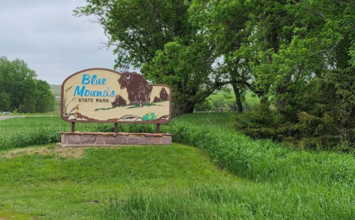 Sign for Blue Mounds State Park surrounded by green grass and trees on a cloudy day.
