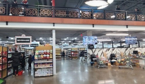Interior of a grocery store with shelves of products, a beverage section, and shoppers browsing.