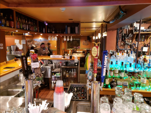 A cozy bar interior with a variety of liquor bottles, a well-stocked counter, and patrons in the background.