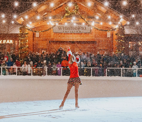 A figure skater performs on an ice rink, surrounded by a festive crowd and twinkling lights in a snowy setting.