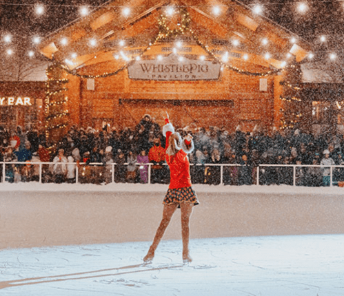 A figure skater performs on an ice rink, surrounded by a festive crowd and twinkling lights in a snowy setting.