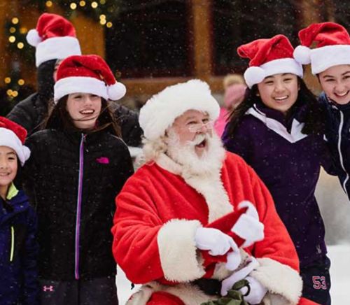 Santa Claus in a red suit sits with a group of children wearing Santa hats, smiling in a snowy outdoor setting.