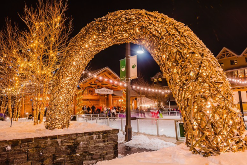 A lit arch made of branches stands in a snowy area, with a cozy lodge and ice skaters in the background at night.