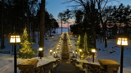 A snowy pathway lined with glowing lamps and trees, leading to a lake at dusk.