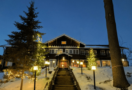 A cozy lodge illuminated with lights, surrounded by snow and trees, under a twilight sky.