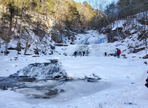 A snowy landscape featuring a frozen waterfall, with people exploring the area and trees in the background.