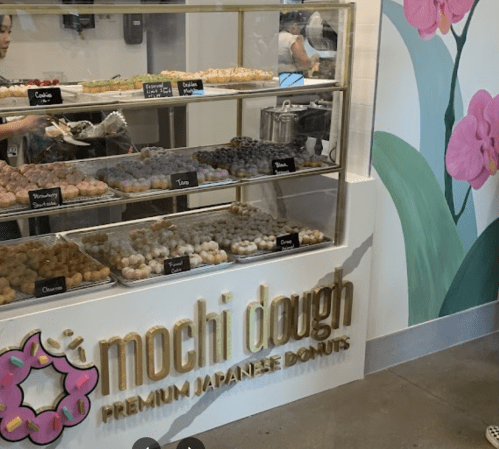 A display of colorful mochi donuts at a shop, with a sign reading "mochi dough" and a floral mural in the background.