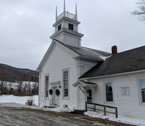 A white church building with a steeple, surrounded by snow and mountains in the background, on a cloudy day.
