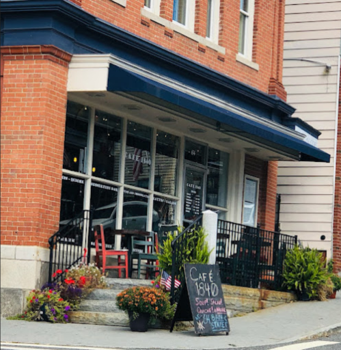 A brick building with a café sign, outdoor seating, and flower planters, featuring a blackboard menu outside.