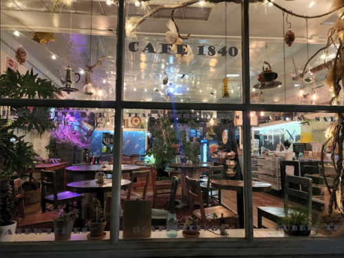 A cozy café interior with plants, colorful lights, and tables visible through a large window at night.