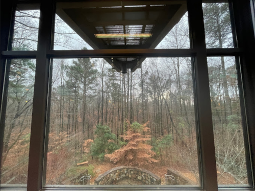 View through a large window showing a forest with bare trees and autumn foliage, under a cloudy sky.
