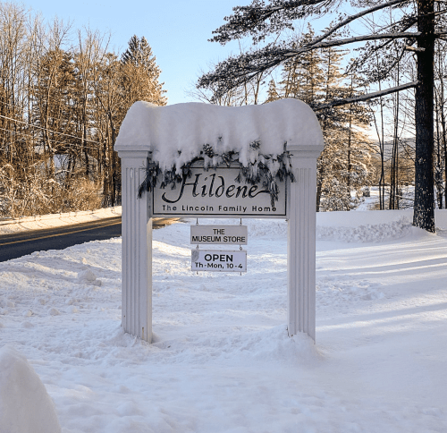 Sign for Hildene, the Lincoln Family Home, covered in snow, with "OPEN" displayed. Winter scene with trees in background.