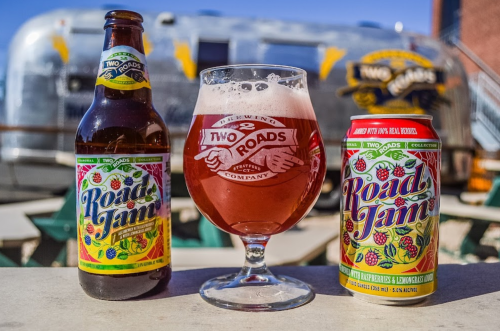 A bottle and can of Two Roads Road Jam beer next to a glass filled with the drink, with an Airstream trailer in the background.