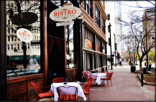 A bistro with outdoor seating and tables set for dining, located on a city street with trees and buildings in the background.