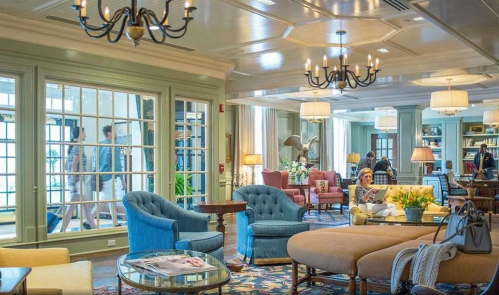 Cozy hotel lobby with elegant seating, chandeliers, and guests walking by large windows.