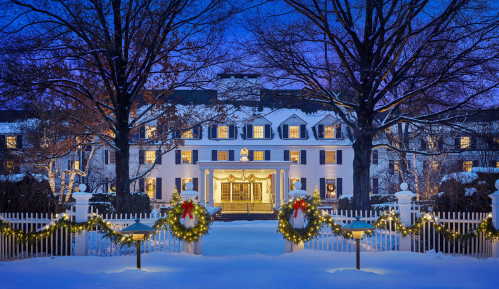 A grand white mansion adorned with holiday decorations, surrounded by snow and lit by warm lights at dusk.