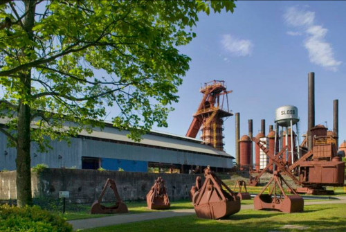 Industrial landscape featuring rusted machinery, tall smokestacks, and a water tower, surrounded by greenery.