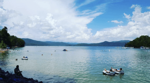 A serene lake scene with people swimming and boating, surrounded by lush greenery and mountains under a blue sky.