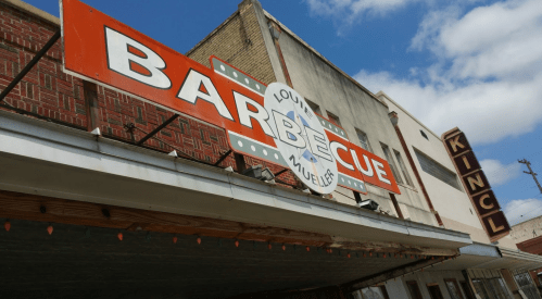 Sign for "Louis Mueller Barbecue" with a blue sky and clouds in the background, featuring colorful lights below.