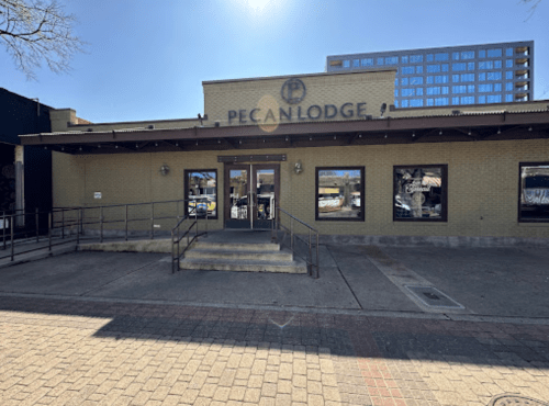 Exterior of Pecan Lodge restaurant with a ramp, large windows, and a clear blue sky in the background.