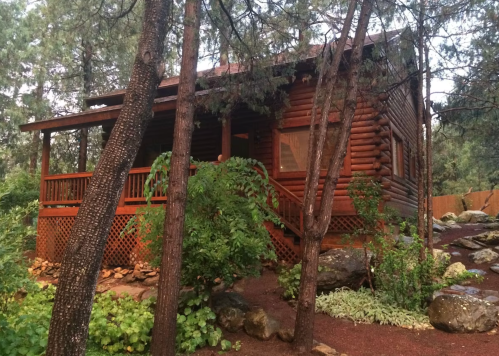 A rustic log cabin surrounded by trees and greenery, with a wooden porch and rocky landscaping.