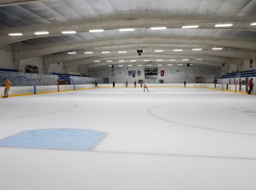 An empty ice rink with skaters practicing, surrounded by bleachers and banners in a well-lit indoor arena.