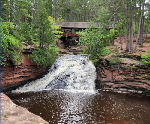 A serene waterfall cascades into a dark pool, surrounded by lush greenery and a wooden structure in the background.