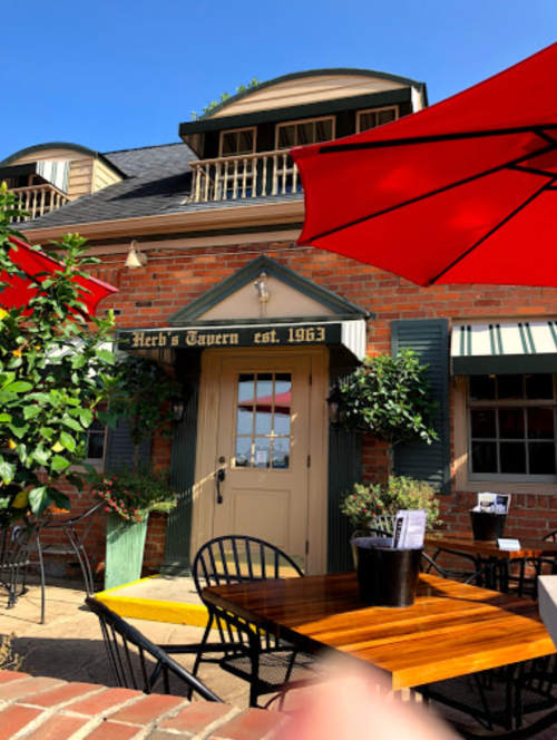 Exterior of Herb's Tavern, established in 1963, featuring outdoor seating and red umbrellas on a sunny day.