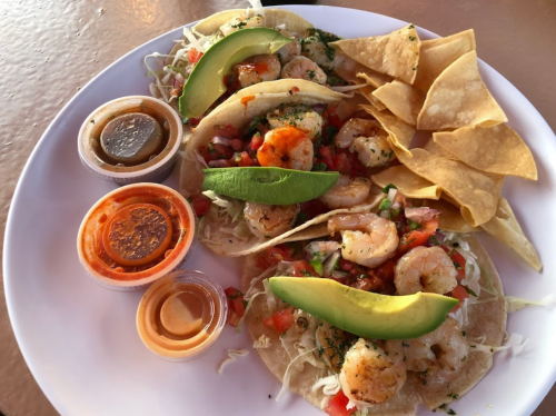 Plate of shrimp tacos topped with avocado, served with tortilla chips and three sauces on the side.