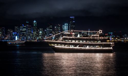 A decorated boat with lights sails on water, with a city skyline illuminated at night in the background.