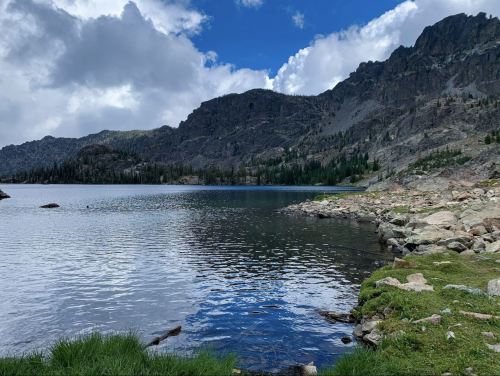 A serene lake surrounded by mountains, with rocky shores and lush greenery under a partly cloudy sky.