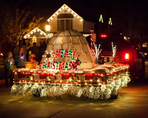 A festive float decorated with lights and an igloo, featuring "Merry Christmas" in colorful letters, during a holiday parade.