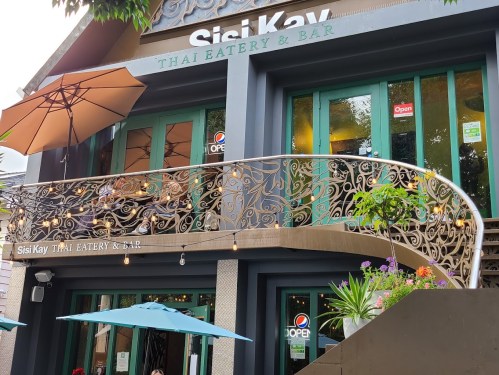 Exterior of Sisi Kay Thai Eatery & Bar, featuring a balcony, green doors, and outdoor seating with umbrellas.